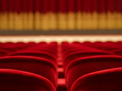 A row of red theatre chairs facing a closed red and gold trimmed theatre curtain