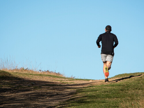 Back of a man running up a hill with grass and a dirt trail